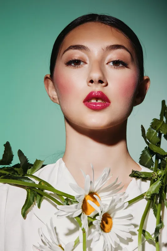 Beauty fashion portrait of a young woman with vibrant red lips and radiant skin. White daisies emphasize natural creative style on a fresh green background. Studio shot with floral accents.