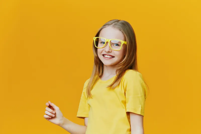Joyful girl in yellow glasses smiling against a vibrant yellow background, showcasing happiness and youthful energy with a playful atmosphere