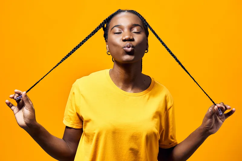 Happy African American woman with braids in a yellow t shirt, playfully posing against a bright yellow background, expressing joy and confidence