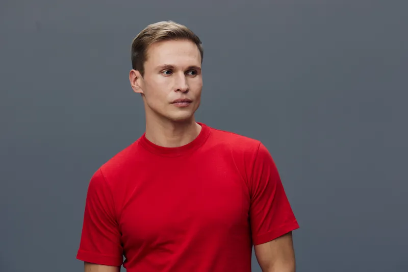 Confident young man with short hair wearing a red t-shirt looks to the side against a plain gray background. Studio portrait, casual style, thoughtful expression, male model, natural colors, minimalis