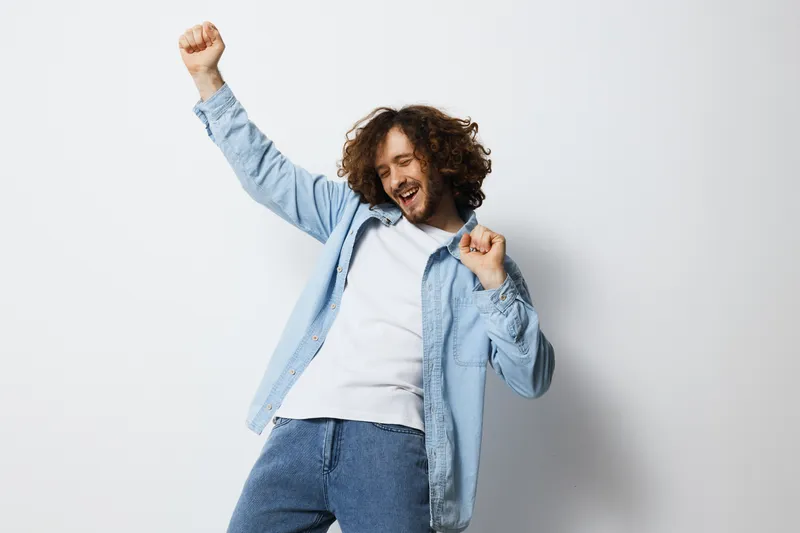 Happy young man dancing joyfully, wearing casual blue shirt and white t shirt, showcasing a positive emotion against a neutral background