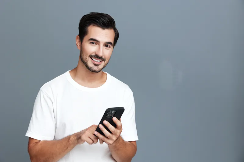Smiling man in casual white t-shirt using mobile phone while standing against a grey background in a studio. Lifestyle and technology concept.