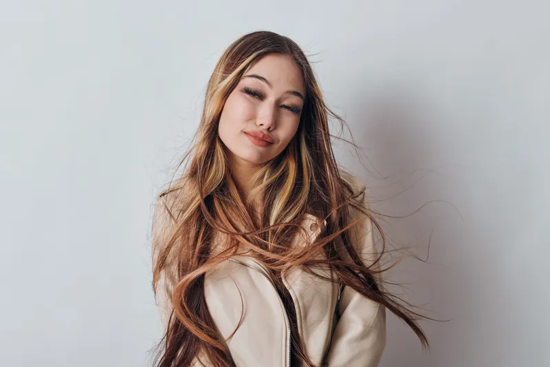 Woman with long hair and closed eyes wearing beige jacket on grey background, beauty portrait with natural makeup and relaxed expression for fashion or lifestyle.