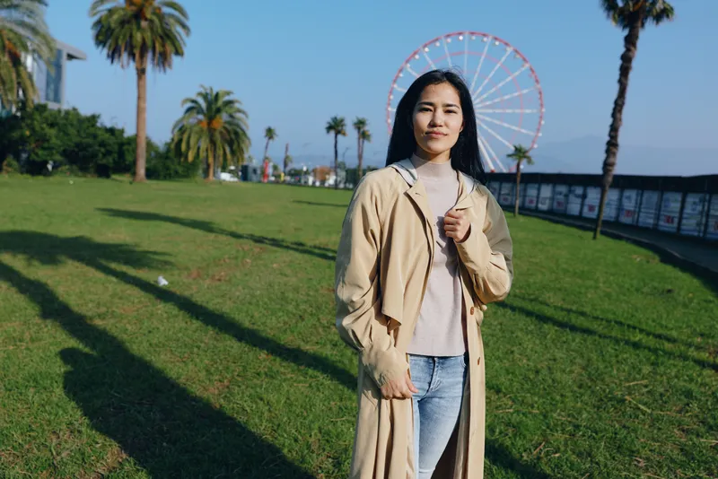 A scenic outdoor scene featuring a woman in a long coat standing on a sunny park lawn with a ferris wheel in the background, palm trees and a boardwalk creating a cheerful seaside ambiance
