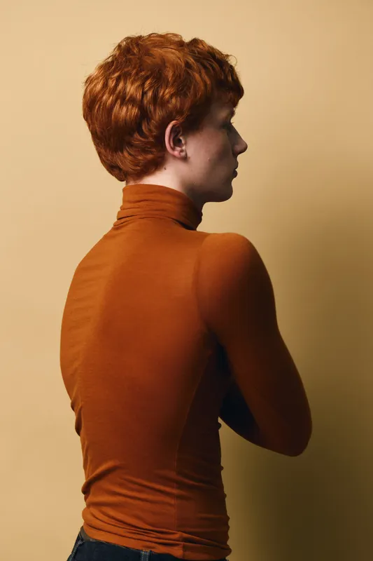 Portrait of young person with red hair and fair skin wearing orange turtleneck, standing sideways against beige background, showing profile and folded arms in studio lighting.