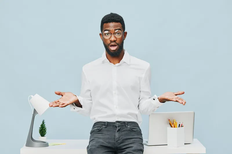Confused man with glasses in white shirt expressing uncertainty while standing behind a desk with a lamp, plant, and office supplies, on plain light blue background. People lifestyle concept.