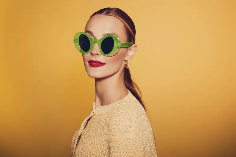 Stylish woman with green round sunglasses and red lipstick poses confidently against yellow background. Fashion portrait with retro vibe and bold color contrast for trendy style concept.