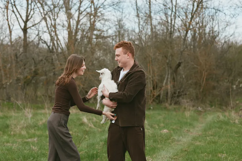 Couple outdoors interact with a joyful goat kid, capturing warm connection and rural lifestyle in a natural field. Soft daylight and candid moment convey happiness, tenderness, and companionship in a