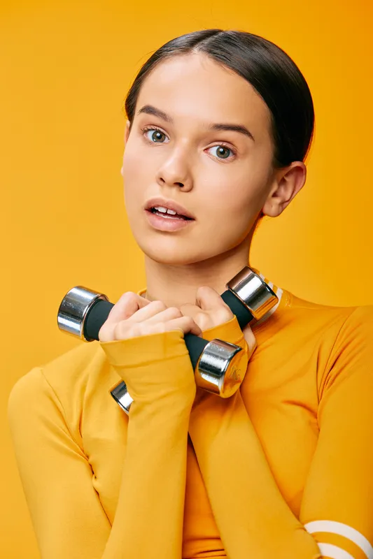 Athletic woman in orange sportswear holds dumbbells near shoulders in a studio with a bold orange backdrop, illustrating fitness training, strength, balance and focus for sport.