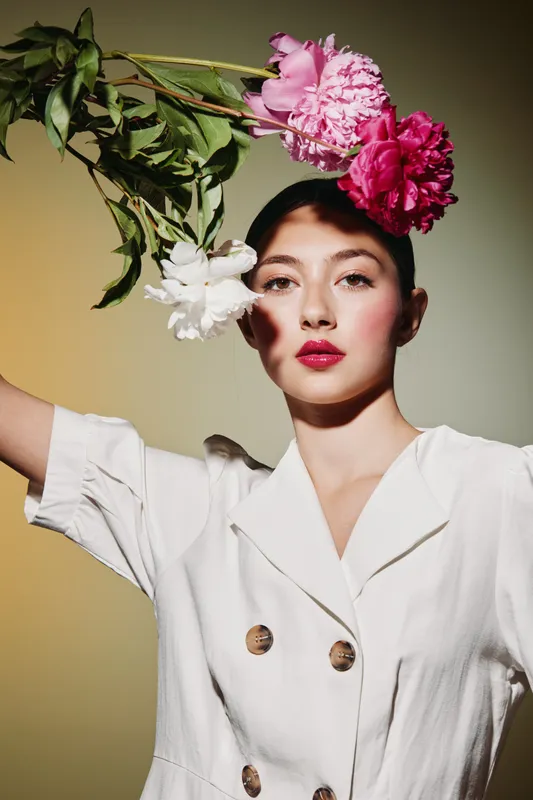 Young woman with bright makeup holds floral branch above head wearing a white buttoned dress, studio shot with dramatic mixed color background beauty, creative fashion concept.
