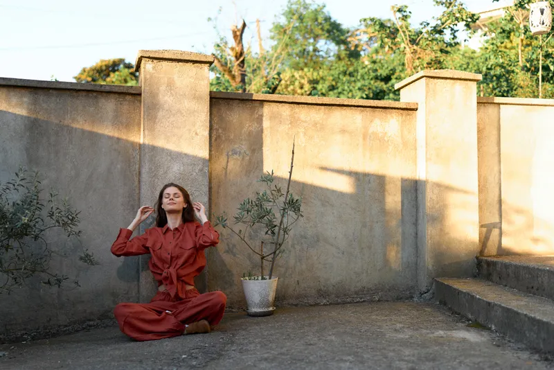A woman in a red dress sits cross legged outdoors beside a weathered wall, eyes closed, hands at temples, conveying stress relief, meditation, and a calm, focused lifestyle moment