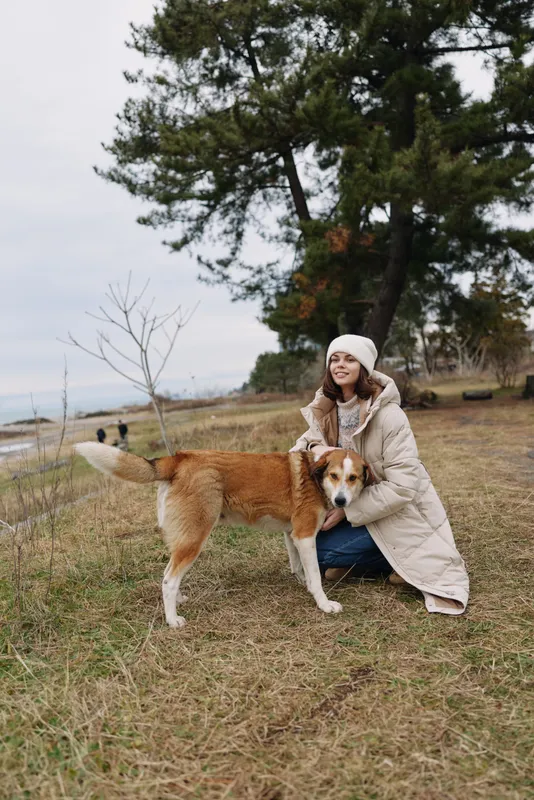 Woman kneels beside a friendly dog in an outdoor park by a lakeside, wearing a warm coat and beanie, enjoying quiet companionship amid trees and calm autumn scenery.