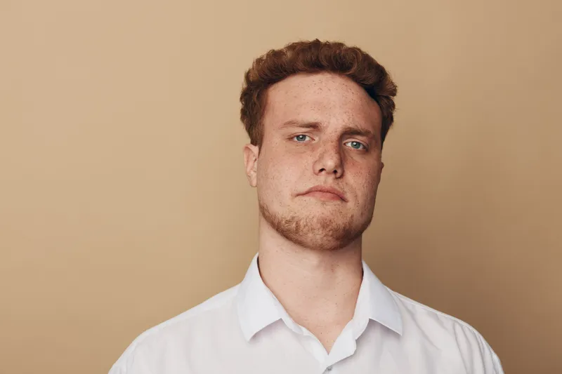 Portrait of serious young man with red hair and beard wearing white shirt against beige background. Concept of confidence, masculinity, and casual style in studio lighting.