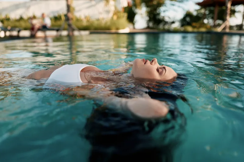 Relaxed woman floating in a sunlit pool, eyes closed, white bikini top, calm expression, warm breeze, natural colors, tranquil outdoor vibe, summer leisure, refreshing water, peaceful getaway