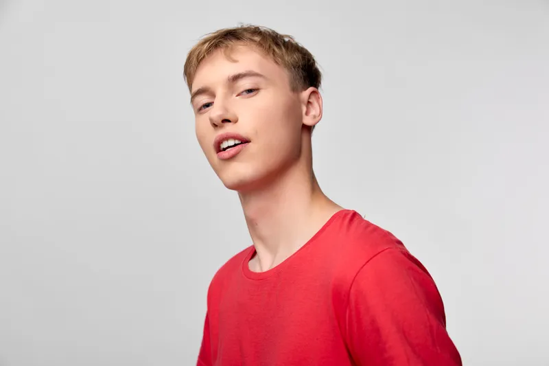 Young man in a red shirt poses in a studio, showcasing casual style and a confident smile with a subtle head tilt, ideal for lifestyle, fashion, or youth-oriented campaigns.