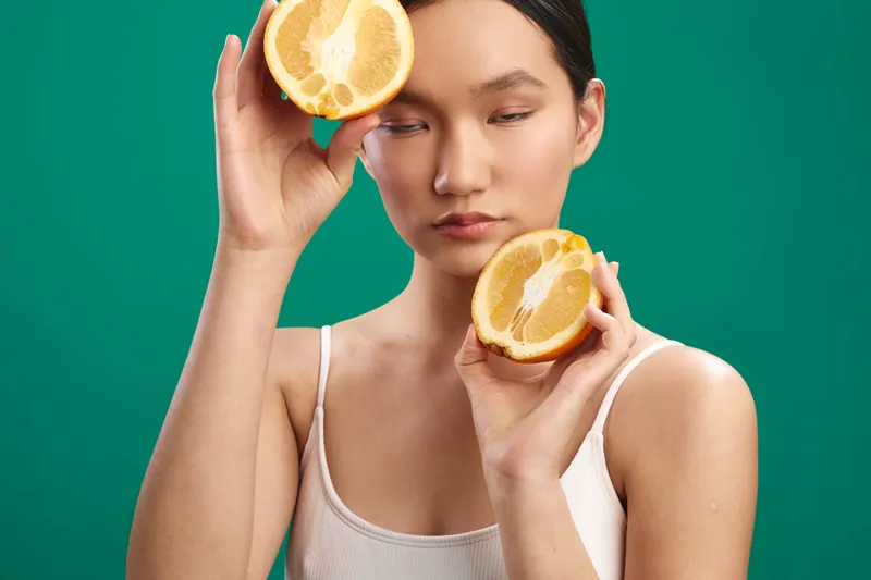 asian woman holds lemon halves near her face in a vibrant studio setting. she gazes softly downward, creating a fresh, citrus inspired portrait with bold color and playful mood.