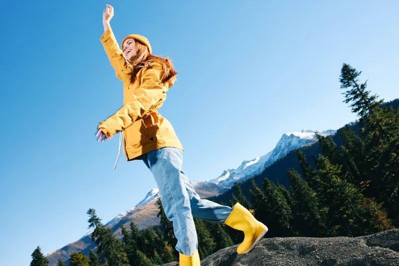 Женщина-туристка с рыжими волосами стоит на горе, подняв руки вверх от счастья, и смотрит на заснеженные горы