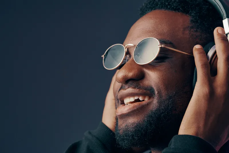 Happy young man enjoying music with headphones, wearing stylish sunglasses against a dark background, vibrant emotions, modern style and contemporary concept