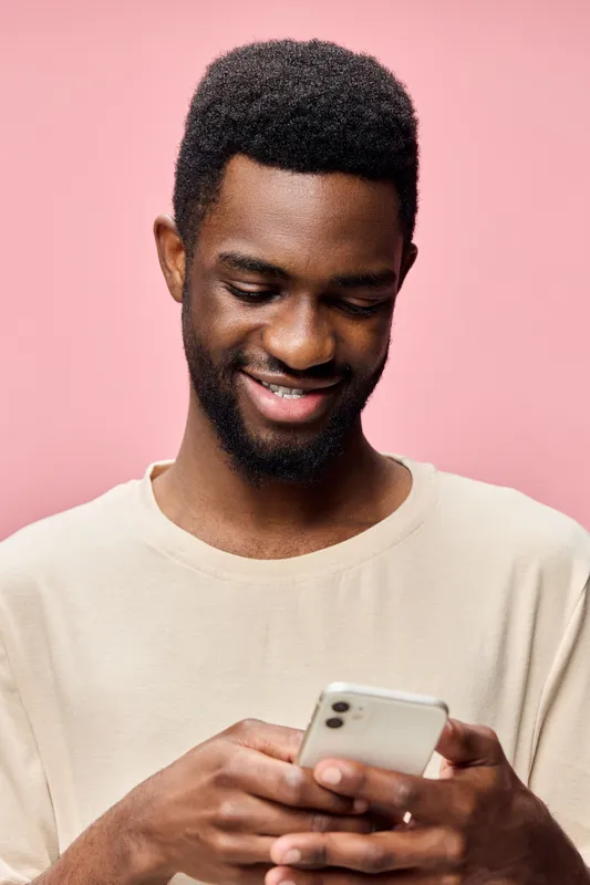 Happy young Black man using smartphone against a pink background, wearing casual light colored clothing, smiling and engaged with mobile technology
