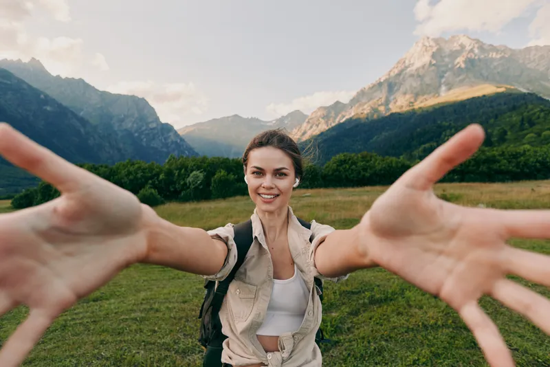 A young woman extends her arms toward the camera in an alpine meadow, inviting connection and adventure amid towering mountains, lush fields, and bright sunlight.
