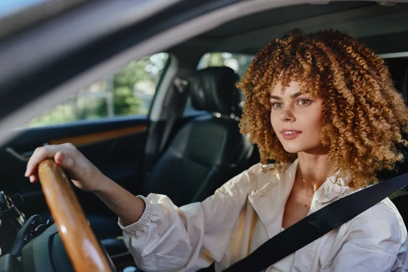 Curly woman driving car with focused expression, wearing white jacket and seatbelt inside modern vehicle, sunny day with natural green background outside window.