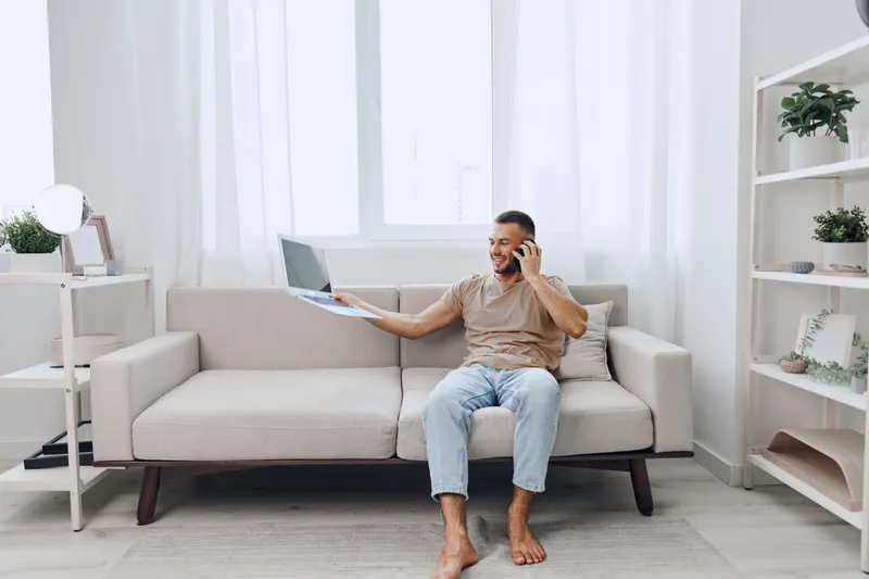 Smiling man sitting on a sofa, engaged in a phone conversation, working remotely with a laptop. Bright, cozy interior with modern decor. Perfect for lifestyle themes.