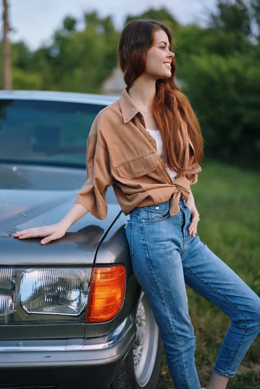 Young woman with long hair in casual outfit poses by a vintage car, exuding confidence and joy in a lush green outdoor setting.
