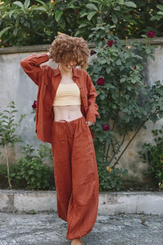 Cheerful woman outdoors, wearing rust orange outfit, standing in a garden, relaxed posture, sunlit moment, natural textures, soft breeze, warm colors, candid lifestyle scene, casual fashion vibe