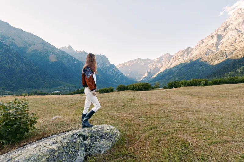 Hiker woman stands on rock with backpack looking over mountains and valley in wide landscape. Outdoor hiking scene, nature adventure, solitude, travel and exploration in alpine meadow.