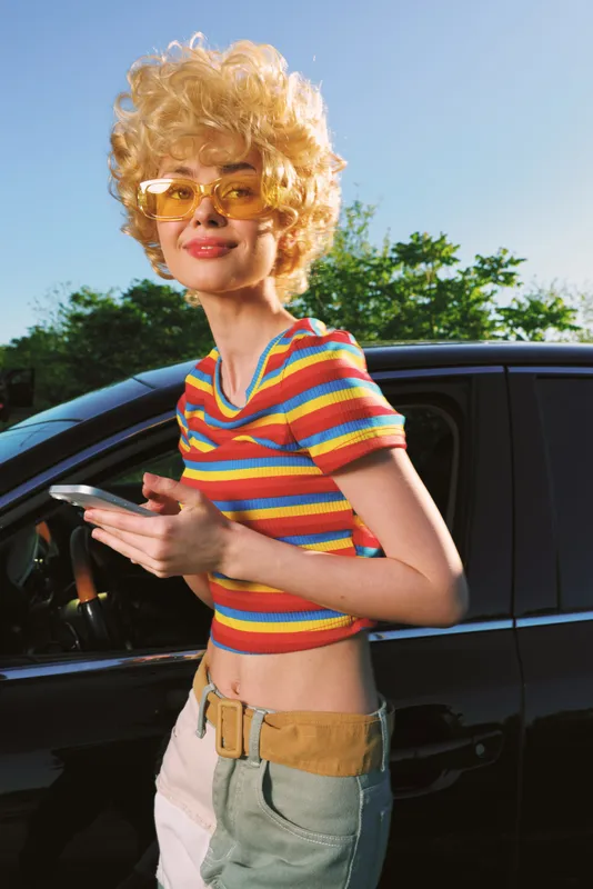 Young woman with retro hairstyle and striped top leans against a car, smiling warmly, captured in warm retro film color for a casual lifestyle portrait.