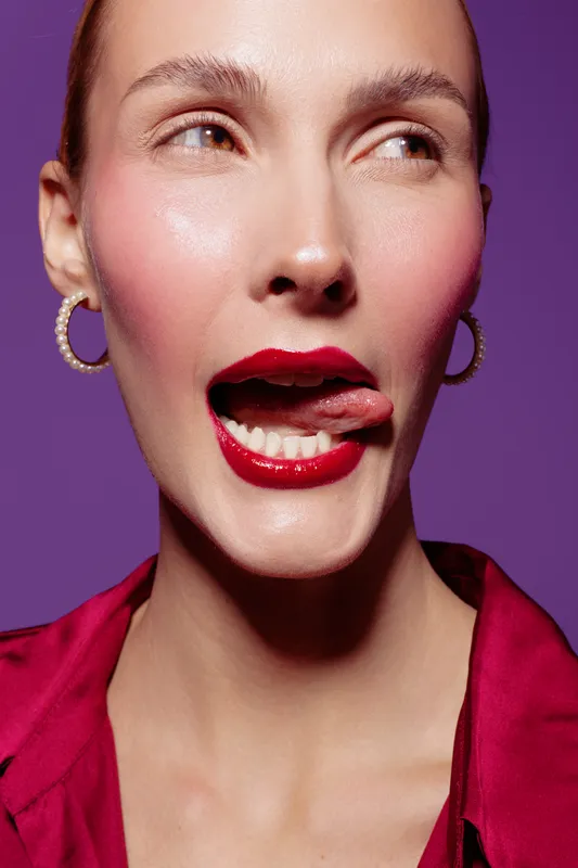 Close-up of a woman with red lipstick and hoop earrings licking her lips against a purple background. Concept of beauty, makeup, fashion, and playful expression.