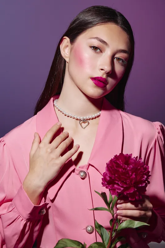 Young woman in pink fashion outfit with vivid makeup holds red peony flower near heart. Soft lighting highlights her pearl necklace against purple background. Beauty creative concept.