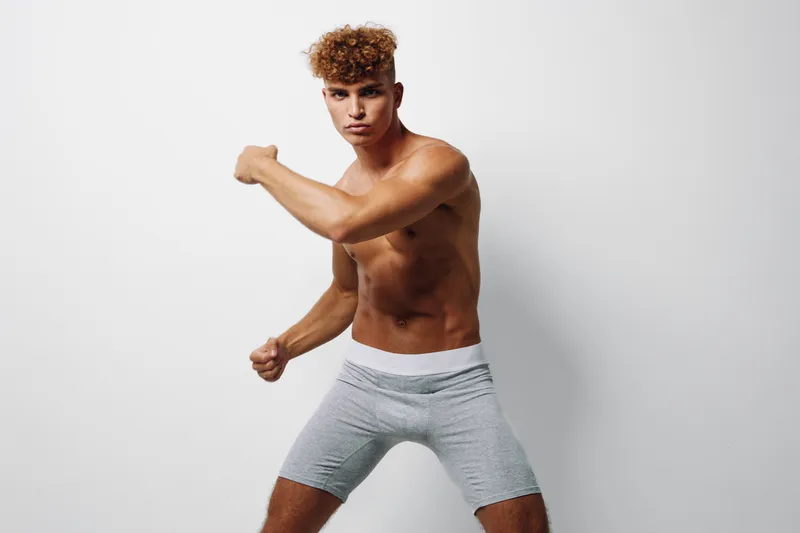 Young athletic man with curly hair posing shirtless in gray shorts against white background, showing muscle definition and fitness in studio lighting.