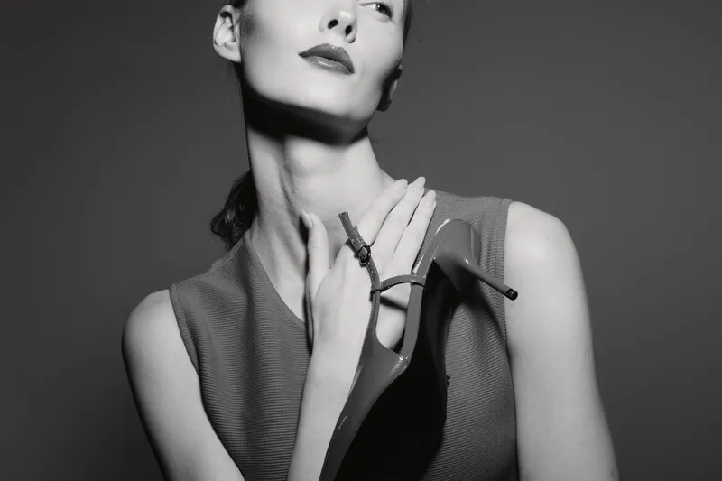 Elegant white woman with pale skin holds high heel shoe near neck in black and white studio portrait.