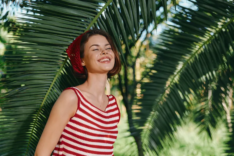 A cheerful young woman wearing a red striped top smiles in a sunny outdoor setting among palm trees, exuding warmth, energy, and a vibrant, carefree lifestyle.