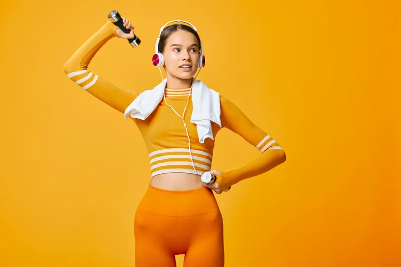 Female fitness portrait of a woman in orange activewear lifting a dumbbell, wearing pink headphones and a white towel, posed confidently against a vibrant yellow studio background.