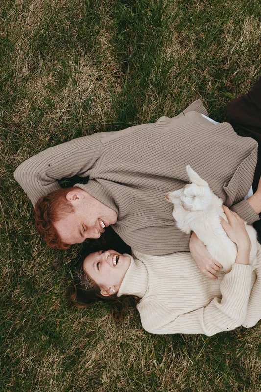 Candid couple outdoors, embracing on grass with a small goat kid nearby, warm sweaters and soft light create a cozy lifestyle moment filled with affection, calm, and natural textures