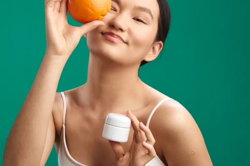 asian woman holding an orange near her eye and a small skincare jar in a bright studio, radiating playful wellness, beauty care, and vibrant mood.