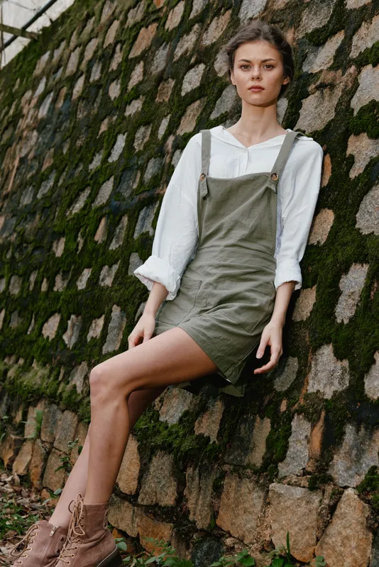 Beautiful young woman with a natural look, wearing a green dress, leaning against a moss covered stone wall in a serene outdoor setting