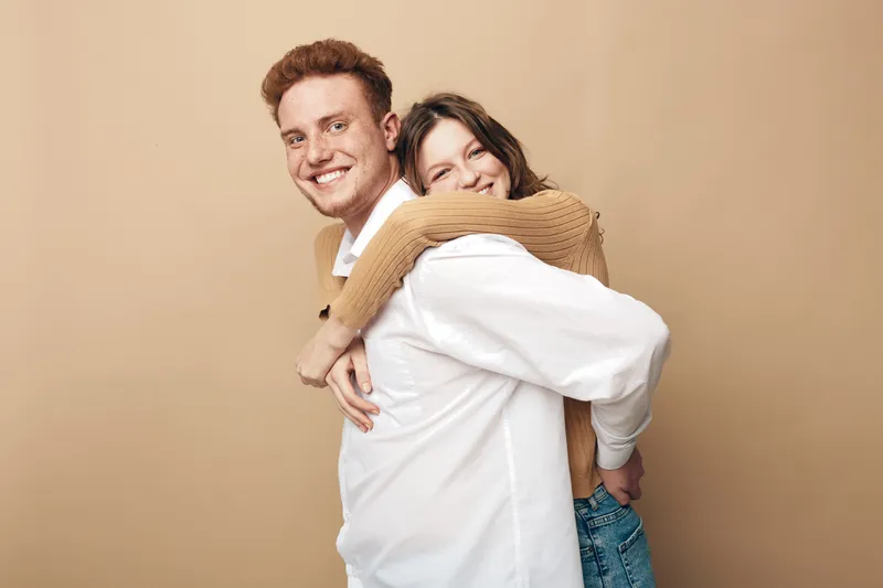 Happy young couple with woman hugging man from behind, both smiling and looking at camera. Casual clothing and warm beige background create friendly and joyful mood.
