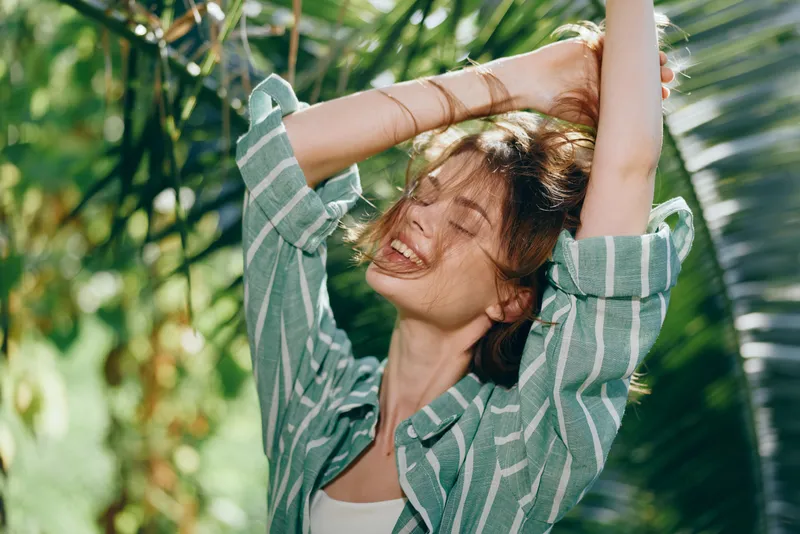 Joyful woman stretching her arms beneath bright sunlight, surrounded by lush greenery. A candid outdoor portrait with warm tones and a relaxed, natural vibe.