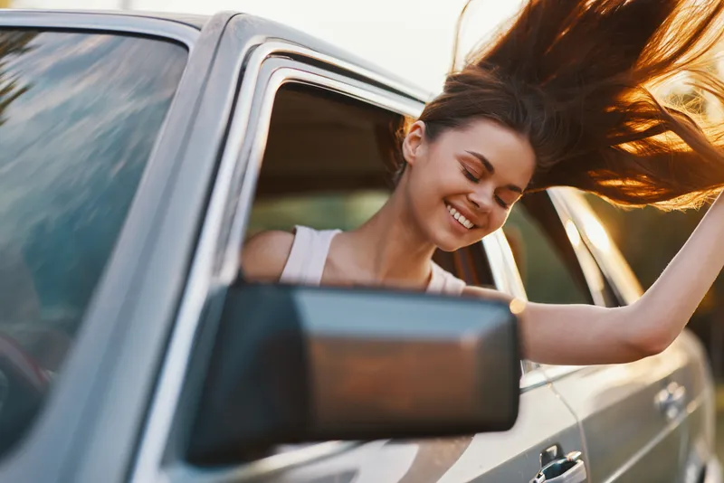 Young woman enjoying a summer drive with her hair blowing in the wind, smiling with happiness while sitting in a car by the roadside during sunset.