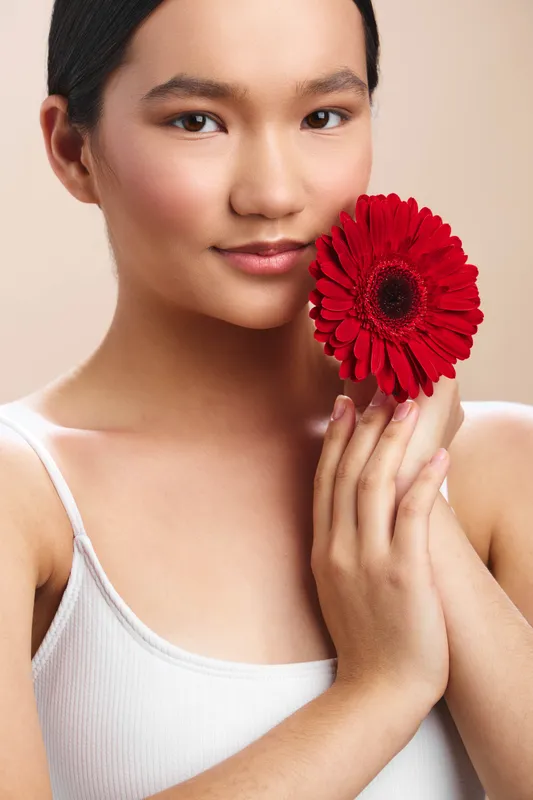 Asian woman wearing a white top holds a vibrant red flower near her face. Soft light highlights smooth skin and a serene expression, creating a gentle beauty portrait.