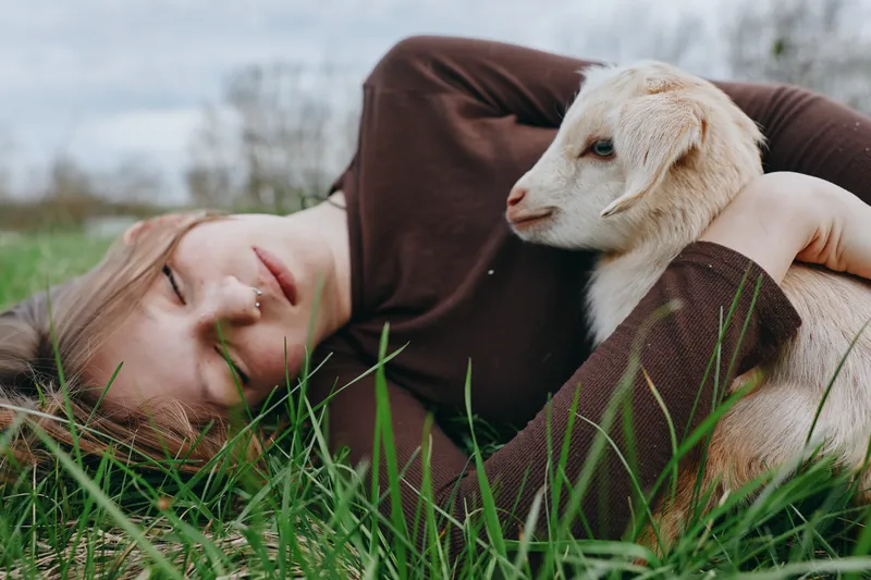 A calm woman lies in the long grass with a gentle goat kid resting beside her, illustrating rural companionship and tenderness in a natural outdoor setting.