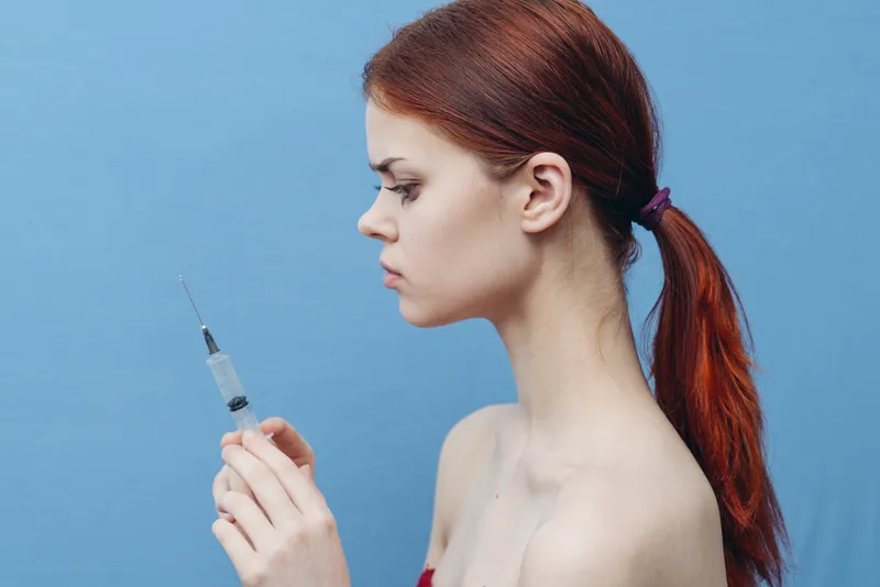 Женщина со шприцем в руках на голубом фоне, инъекция для омоложения ботоксом, вид сбоку