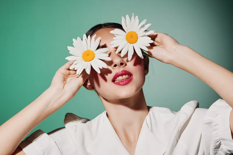 Creative beauty portrait of a young woman with fresh makeup holding white daisies over her eyes on green background. Concept of fashion and artistic expression.