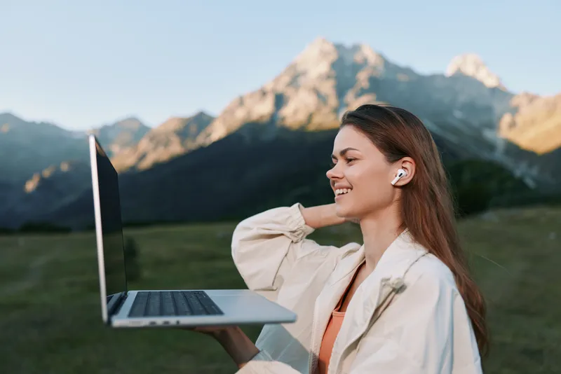 A cheerful professional woman is seen using a laptop outdoors in a scenic setting. She smiles, appears engaged, and focused, creating a positive and dynamic work atmosphere under a clear sky.