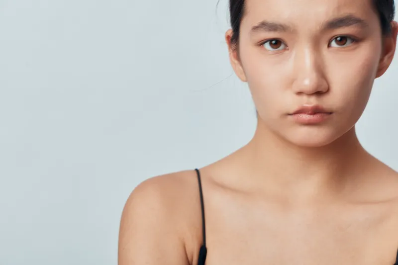 Portrait of young woman with serious expression wearing black top on plain background. Asian female model with natural skin, minimal makeup, and focused gaze in closeup.
