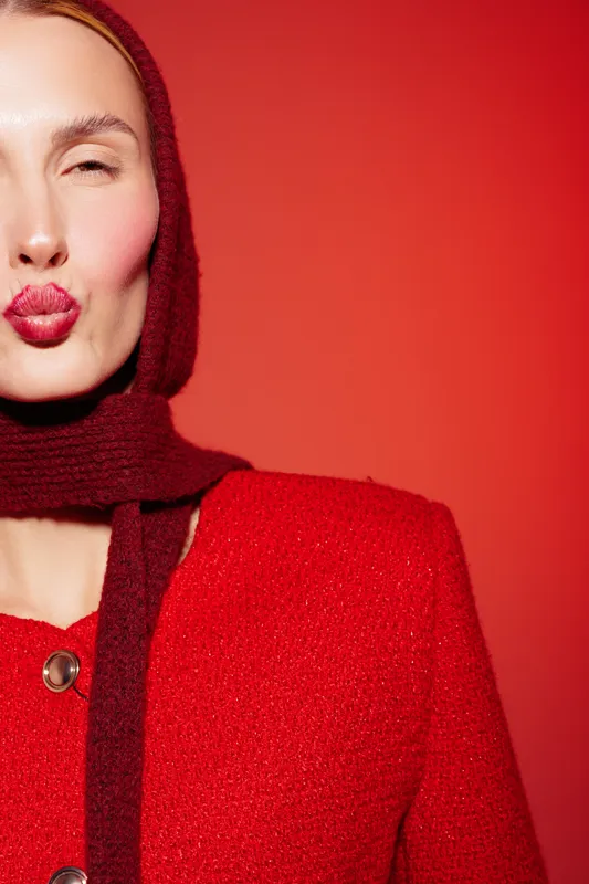 Close-up of woman in red coat and scarf making a kiss face on red background. Fashion portrait with warm tones and playful expression, concept for style and winter clothing.