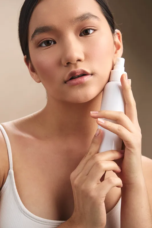 Asian woman applies skincare product in a clean, minimalist space with soft lighting, conveying calm selfcare and a gentle, contemplative beauty routine.